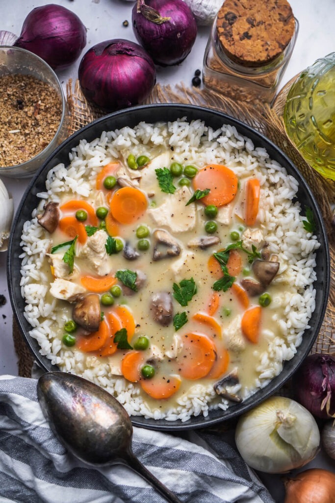 Ein Teller gefüllt mit Reis, oben drauf eine große Portion Hühnerfrikassee it Erbsen, Karotten und Champignons.