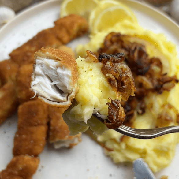 Eine Gabel mit einem Stück Fischstäbchen, Kartoffelpüree und gebratenen Zwiebelringen